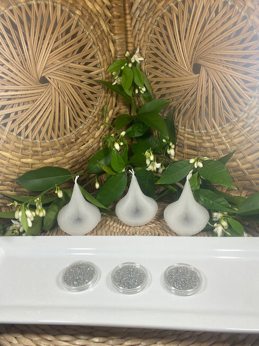 The White Trio Destiny Decision Candles set and the Destiny Sparkles on a white plate, placed on a natural wicker table with green leaves and tiny white Valencia orange blossoms in the background.