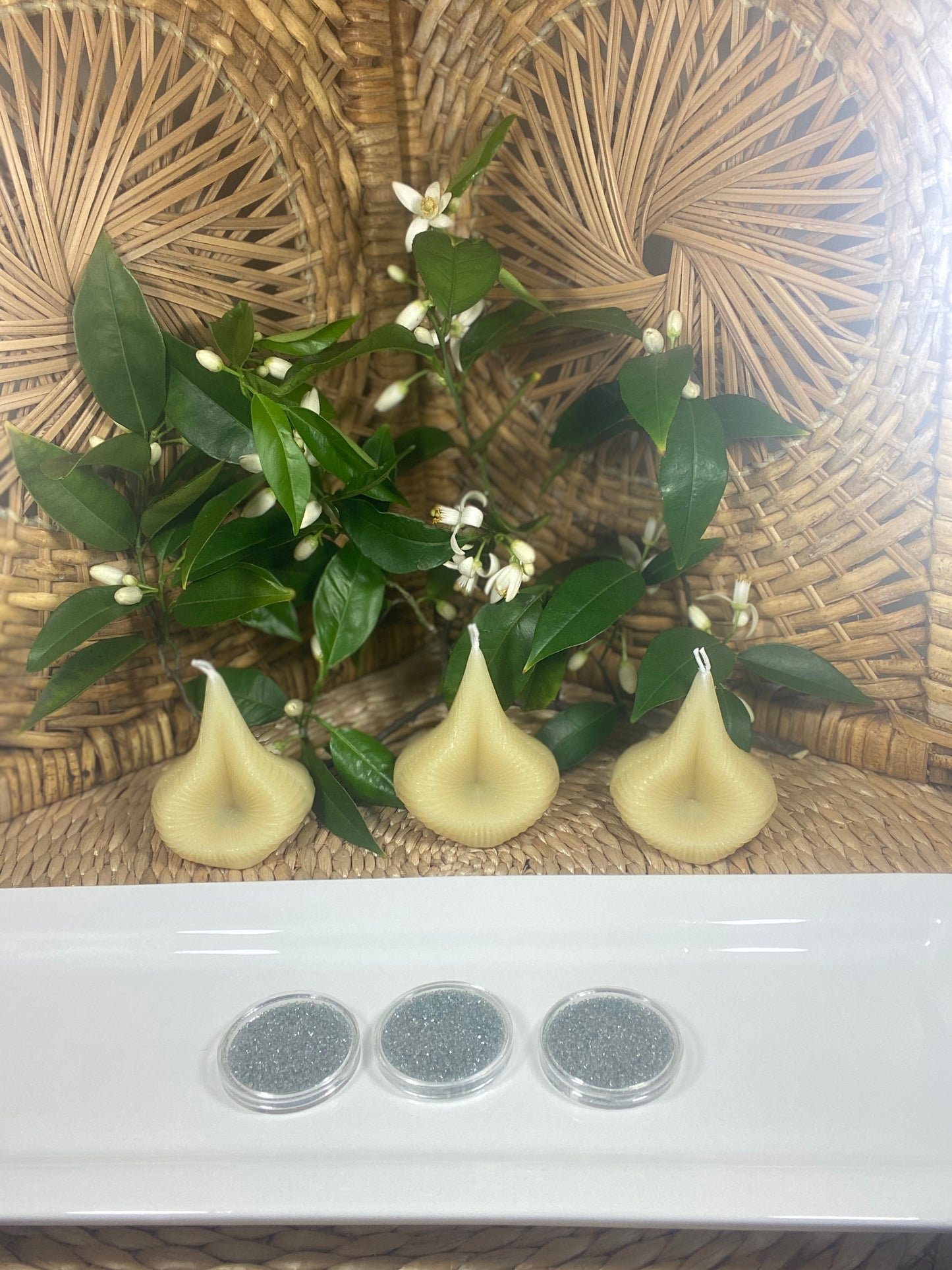 The Natural Golden Beeswax Trio Destiny Decision Candles set and the Destiny Sparkles on a white plate, placed on a natural wicker table with green leaves and tiny white Valencia orange blossoms in the background.